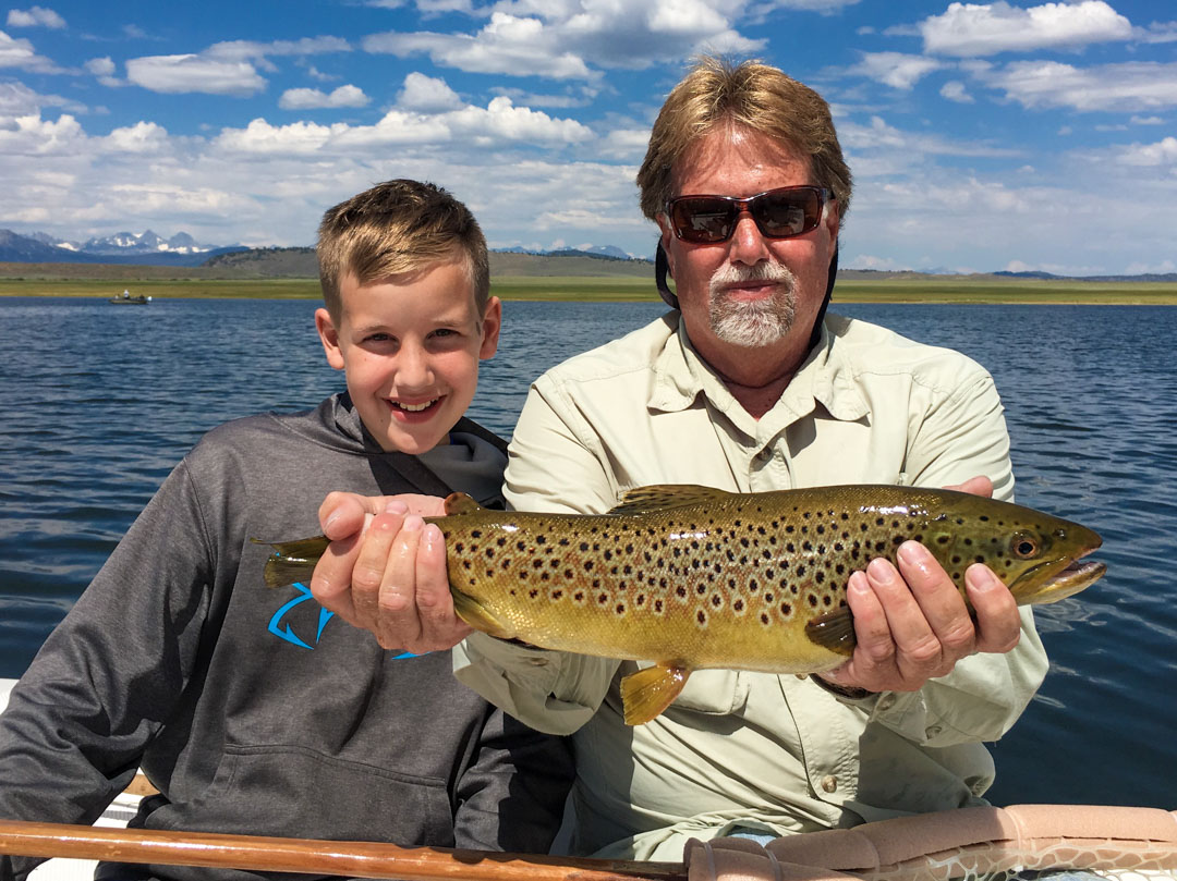  A man and a kid in a boat holding a large brown trout on a lake.