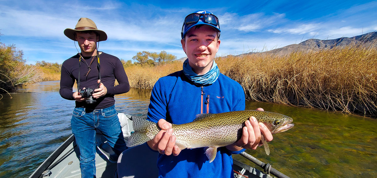 A fly fisherman holding a rainbow trout while standing in a drift boat on the Lower Owens River with a young male photographer in the background..