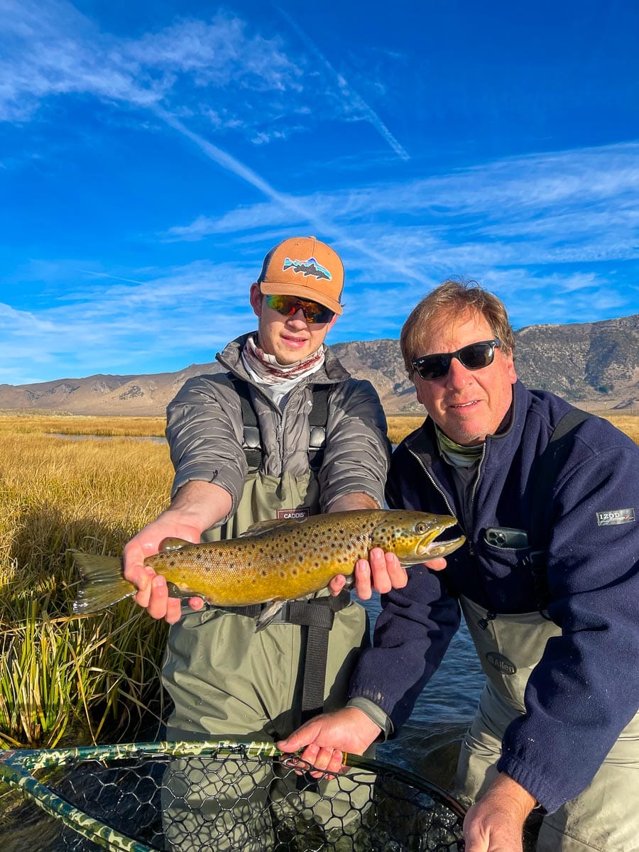 A young fly fisherman holds a brown trout in spawning colors in the fall from the Upper Owens River while his dad assists holding the net.