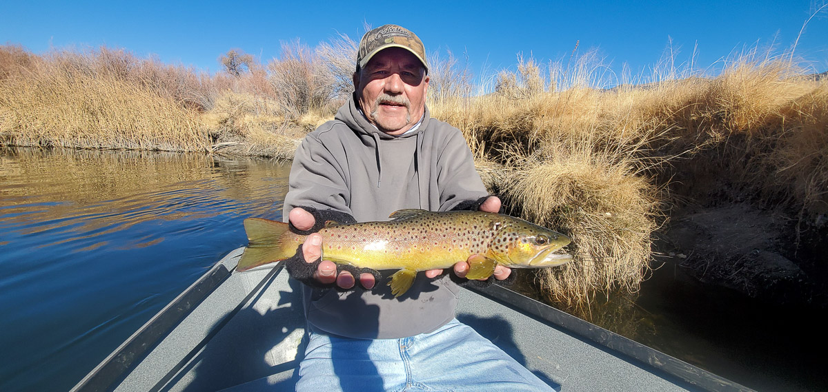 A fly fisherman sitting in a drift boat and holding a brown trout from the Lower Owens River.
