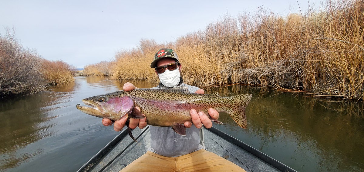 A fly fisherman sitting in a drift boat and holding a rainbow trout from the Lower Owens River.