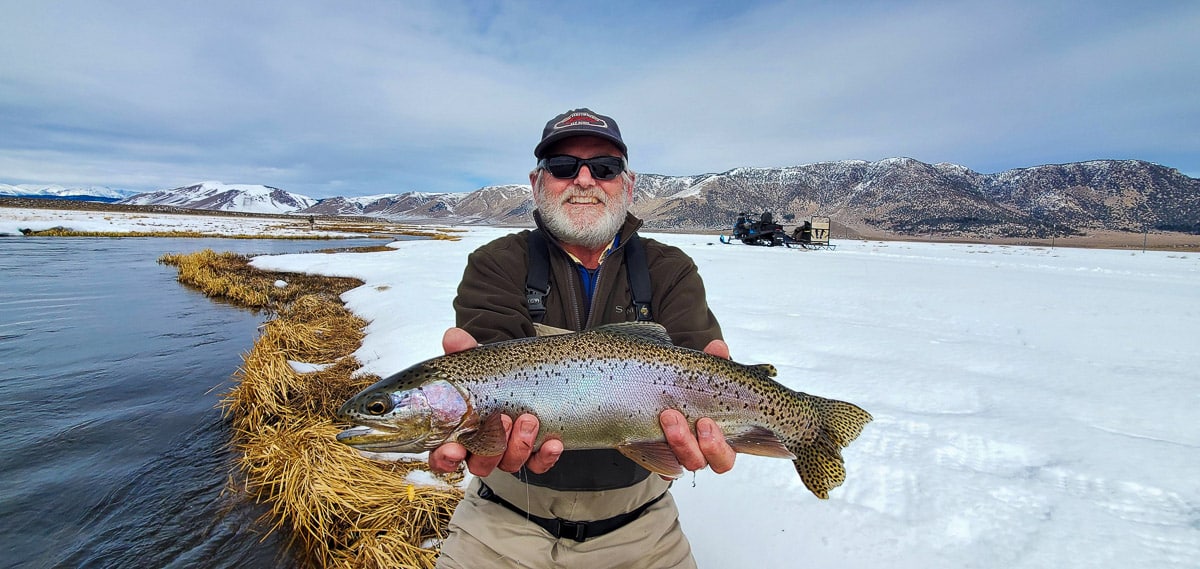 A fly fisherman holding a rainbow trout in spawning colors from the Upper Owens River in the snow.