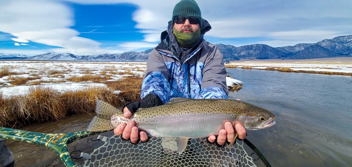 A fly fisherman holding a rainbow trout in spawning colors from the Upper Owens River in the snow.