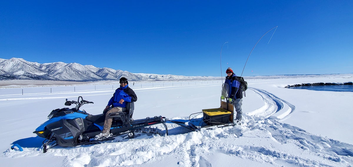 A blue snowmobile carrying a fly fisherman with another fly fisherman on a dog sled trailer.