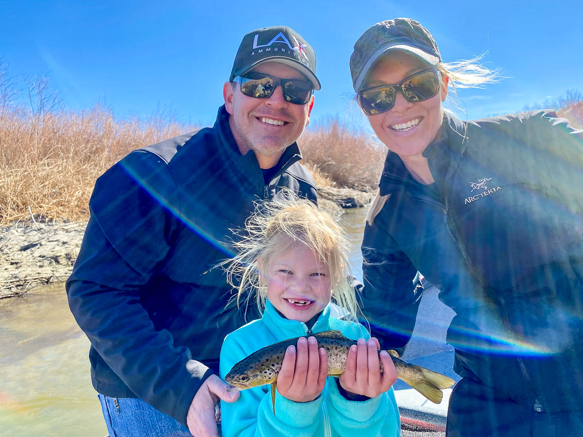 A young family of fly fishers smiling with their 6 year old daughter who is holding a wild brown trout caught from a river.