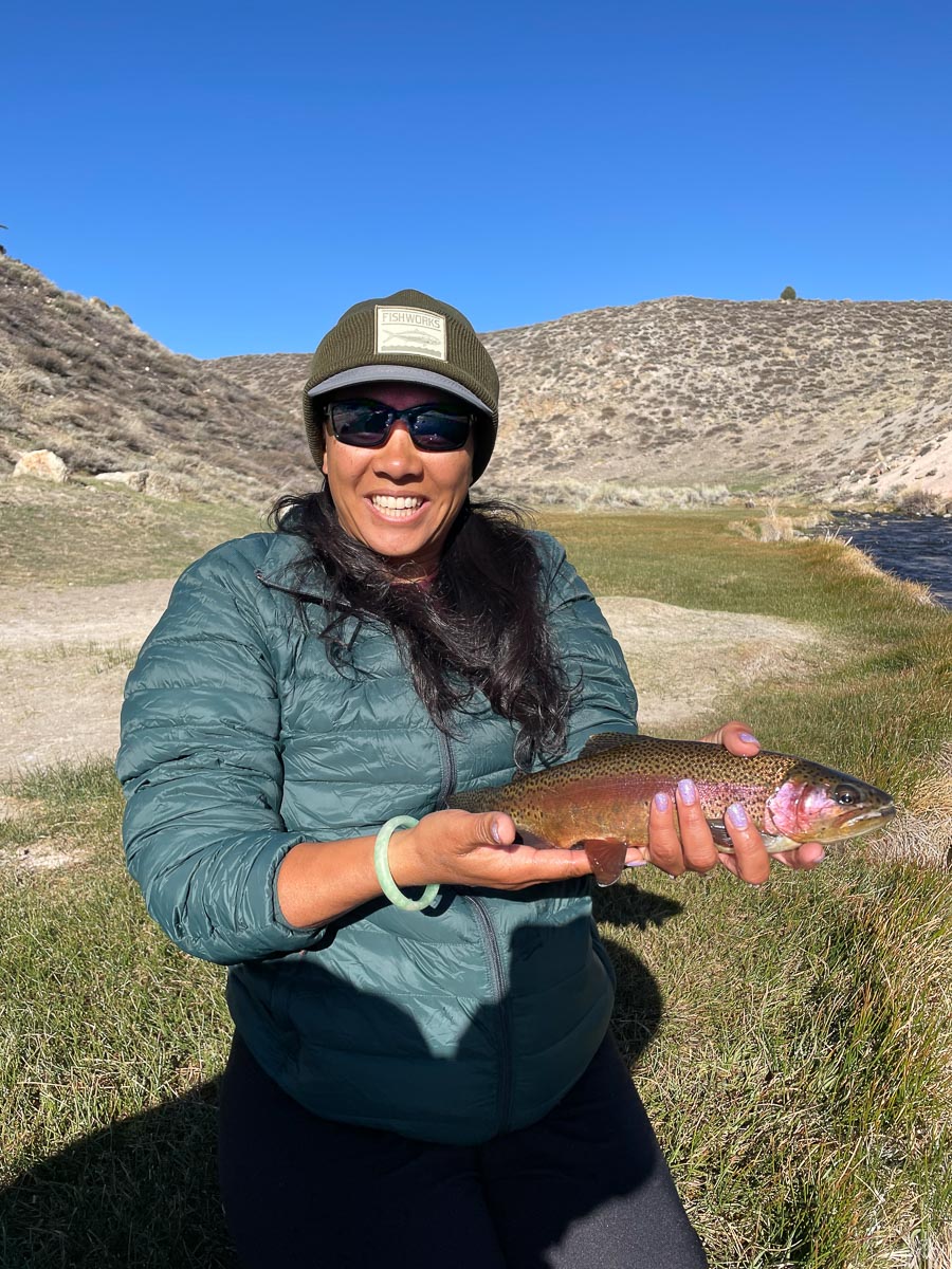 A female fly fishing angler holding a rainbow trout while kneeling on the grassy bank of a river.