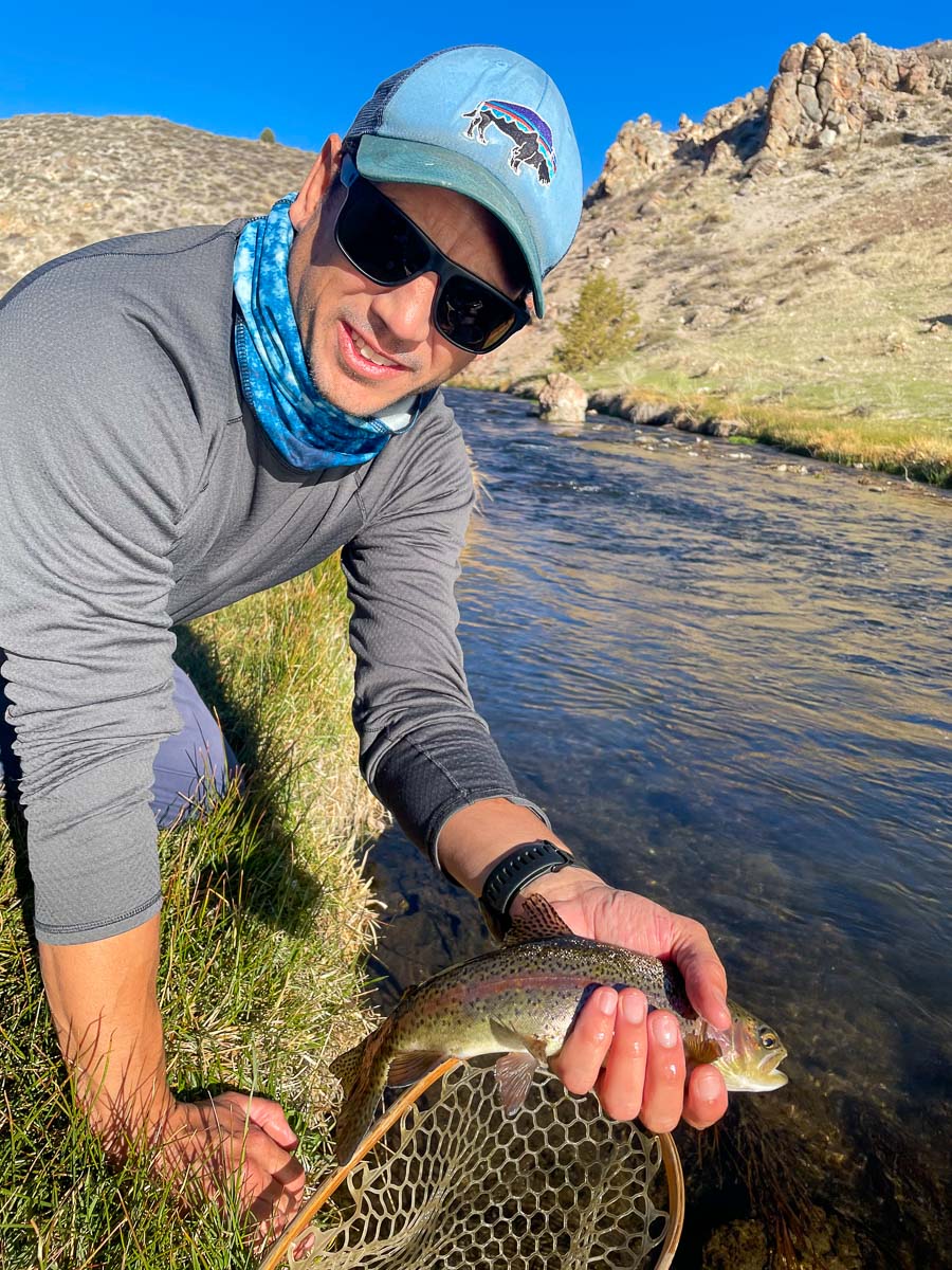 A fly fisherman with a light blue hat kneeling on a grassy river bank holding a rainbow trout in one hand.