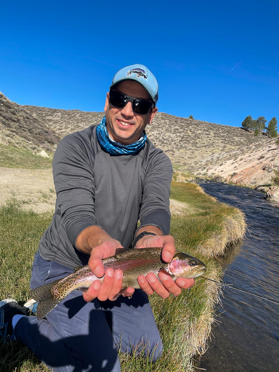 A fly fisherman with a light blue hat kneeling on a grassy river bank holding a rainbow trout in one hand.