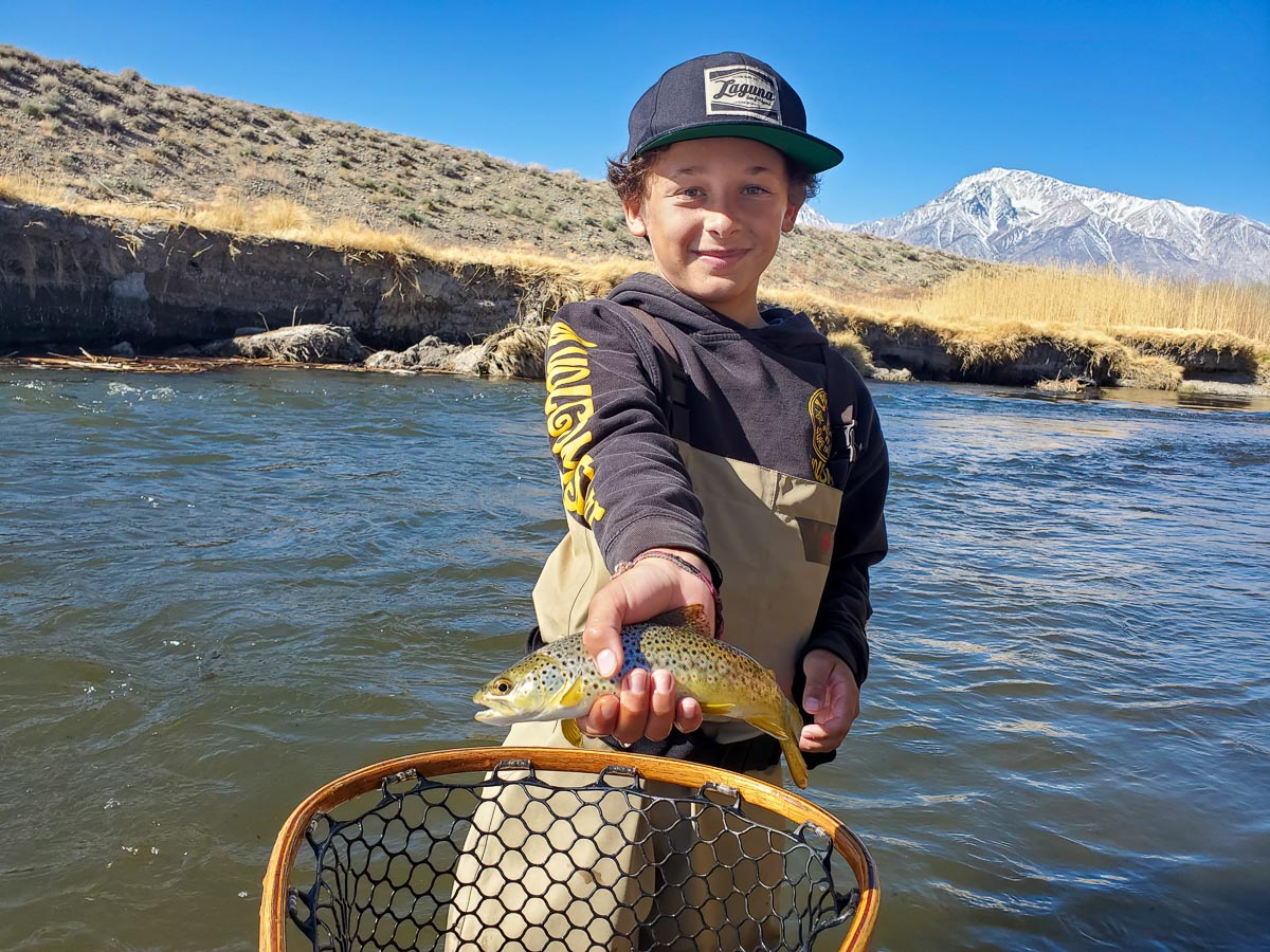 A young fly fisherman standing in a river holding a brown trout.