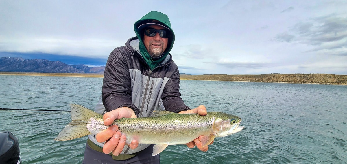 A green hooded fly fisherman holding a fat rainbow trout on a lake.