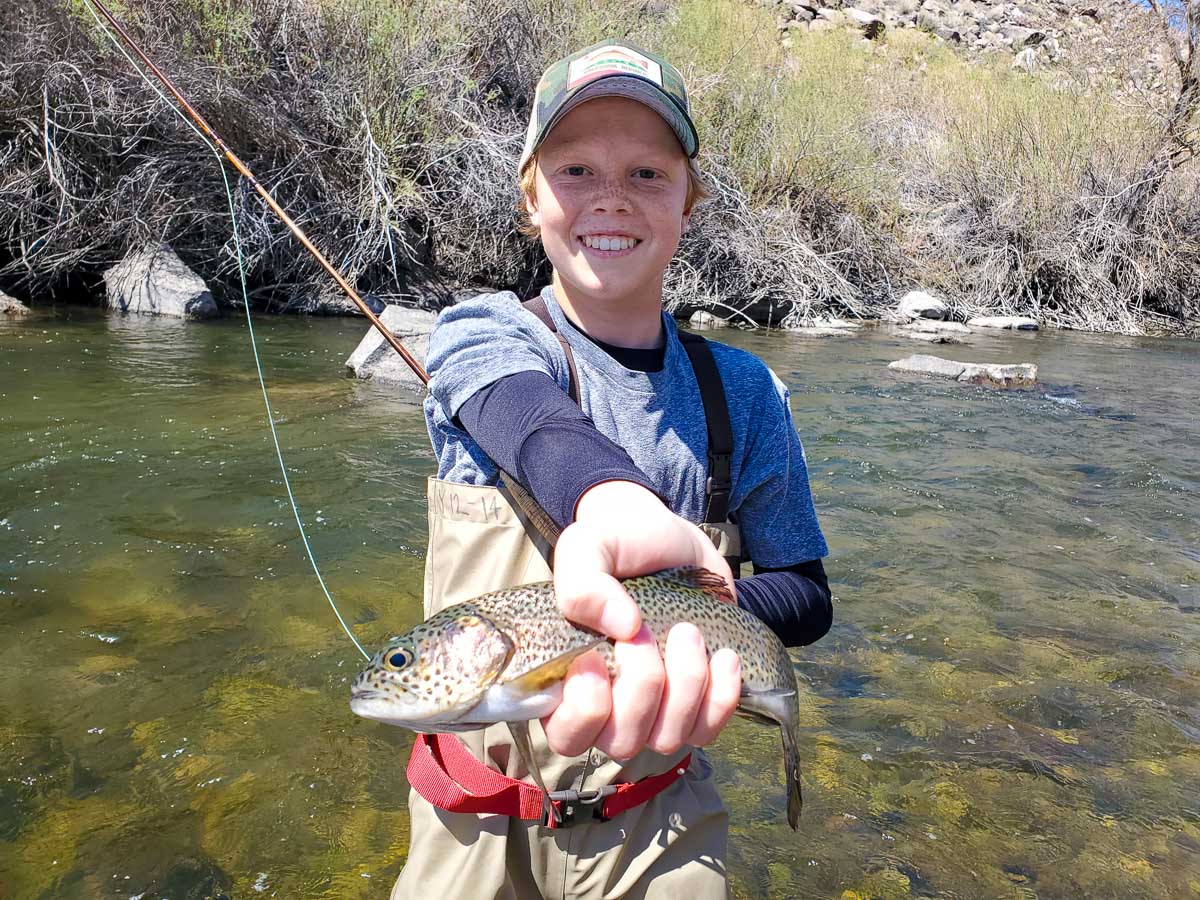 A fly fisherman youth holding a rainbow trout while standing in a river.
