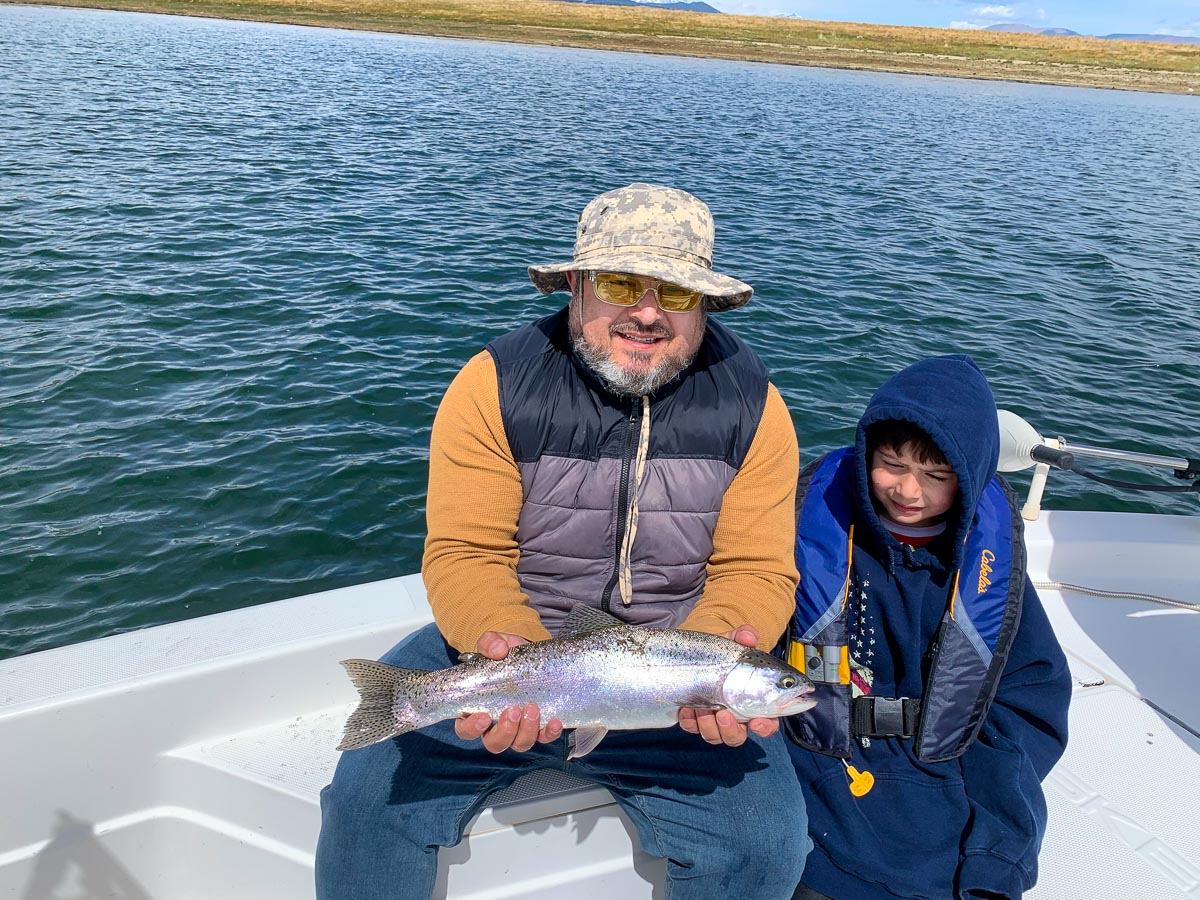 A fly fisherman holding a large rainbow trout in a boat on a lake with his young son by his side.