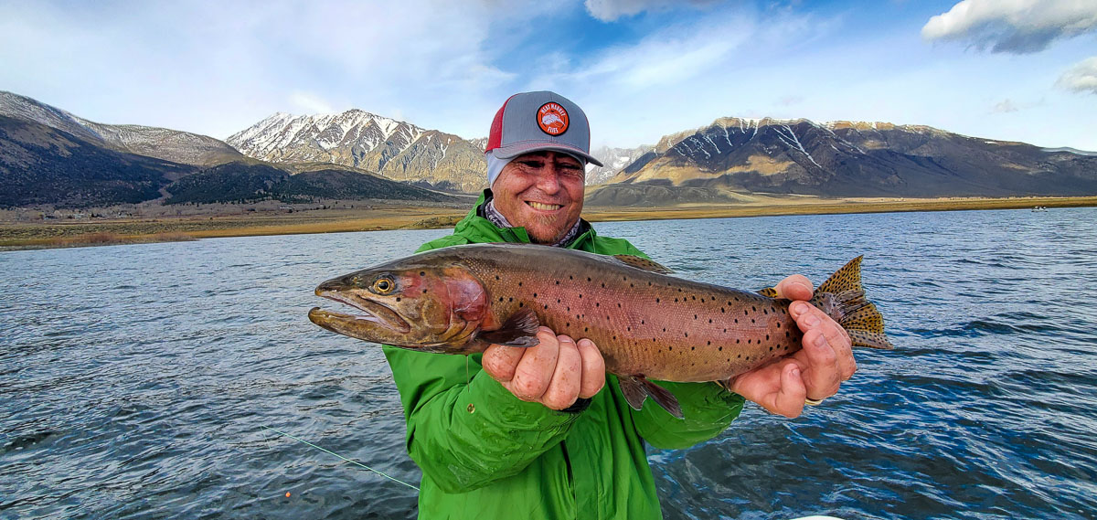 A fly fisherman wearing a grey baseball cap holding a large cutthroat trout on a lake.
