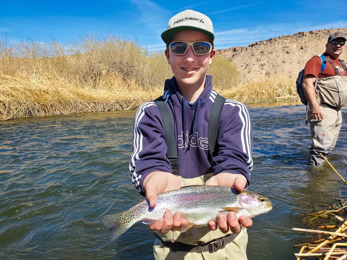 A young fly fisherman standing in a river holding a rainbow trout with a man in the background.