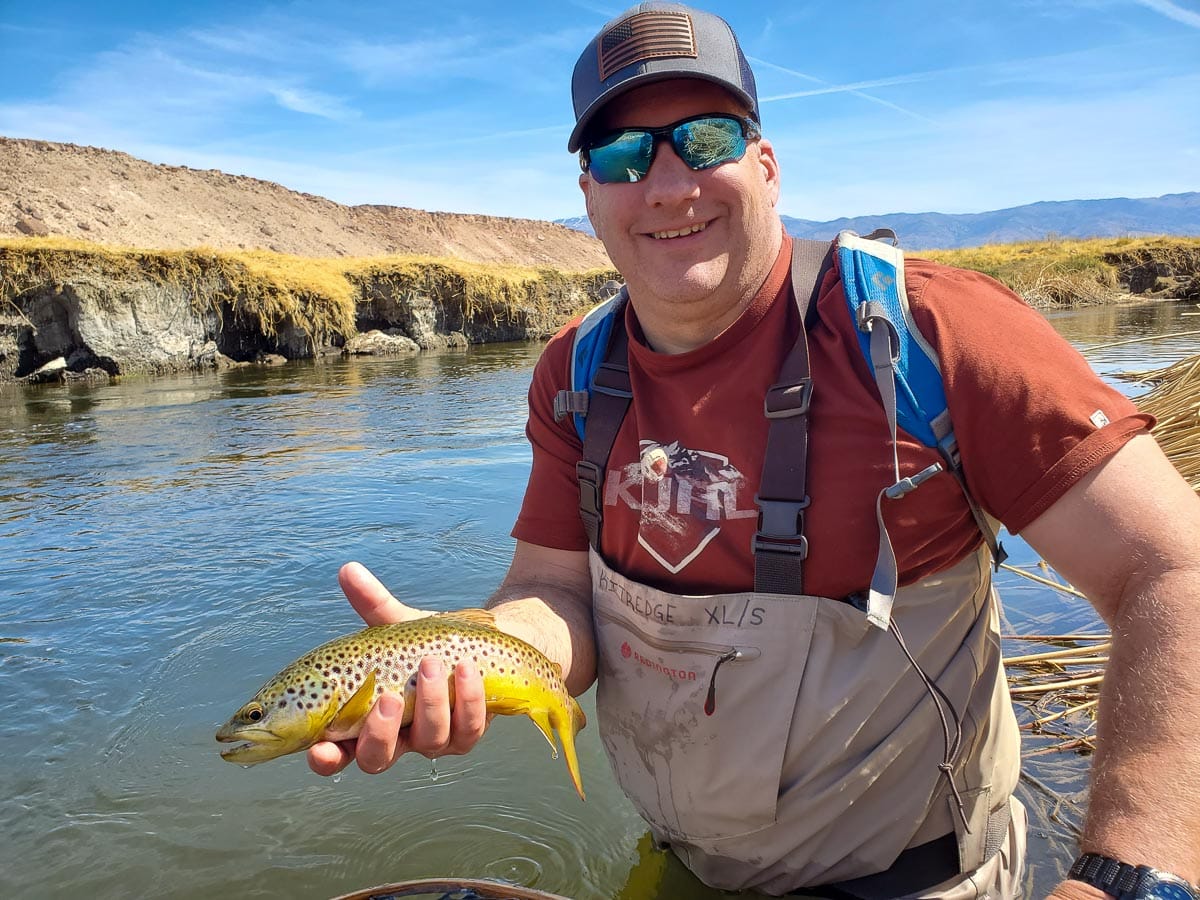 A fly fisherman standing in the river holding a brown trout.