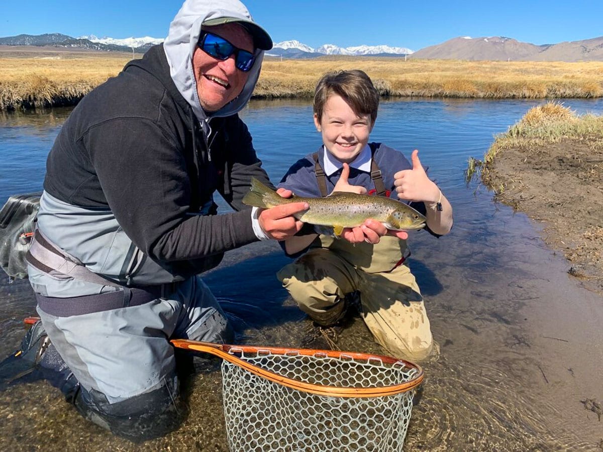 A young fly fisherman kneeling in the water with a hooded man wearing waders holding a brown trout.