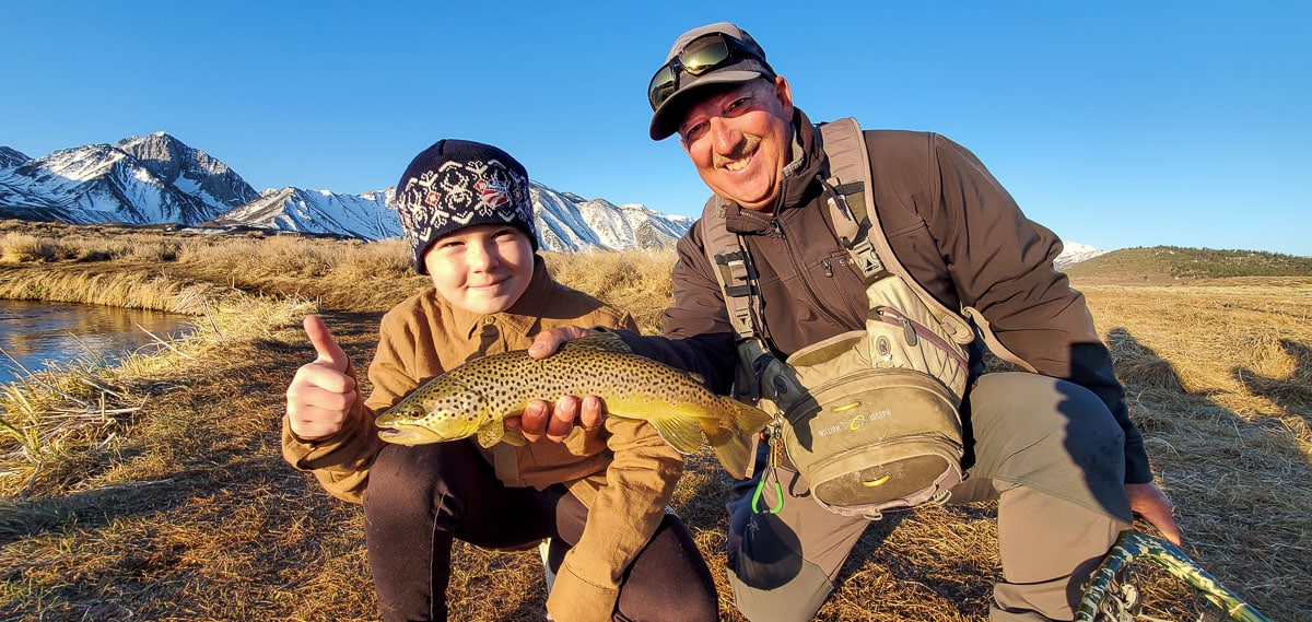 A young fly fisherman kid kneeling next to a man who is holding a brown trout with snowy mountains in the background.