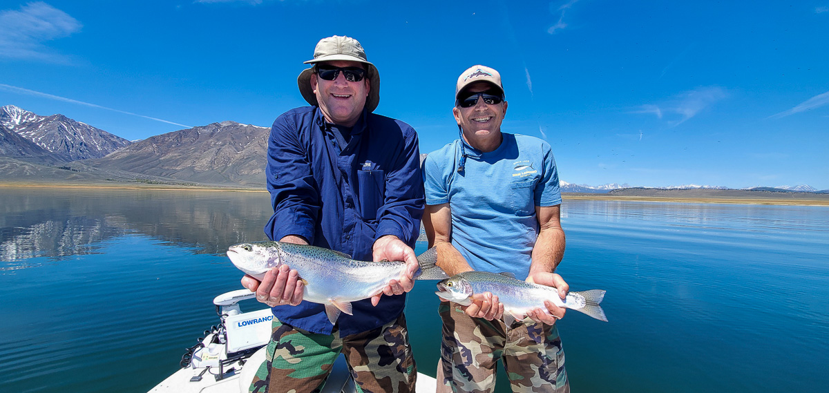 A pair of fly fishermen holding a pair of healthy and shiny rainbow trout in a boat on a mountain lake.