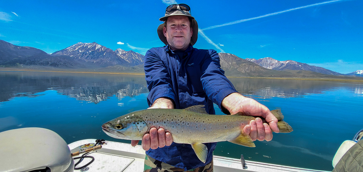 A fly fisherman with a navy blue long sleeve shirt holding a large brown trout in a boat on a lake.