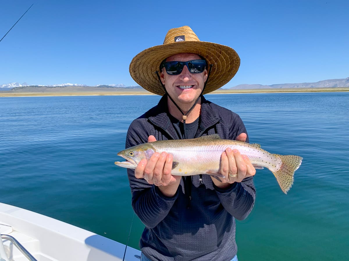 A fly fisherman with a wide-brim hat holding a cutthroat trout on a lake. 