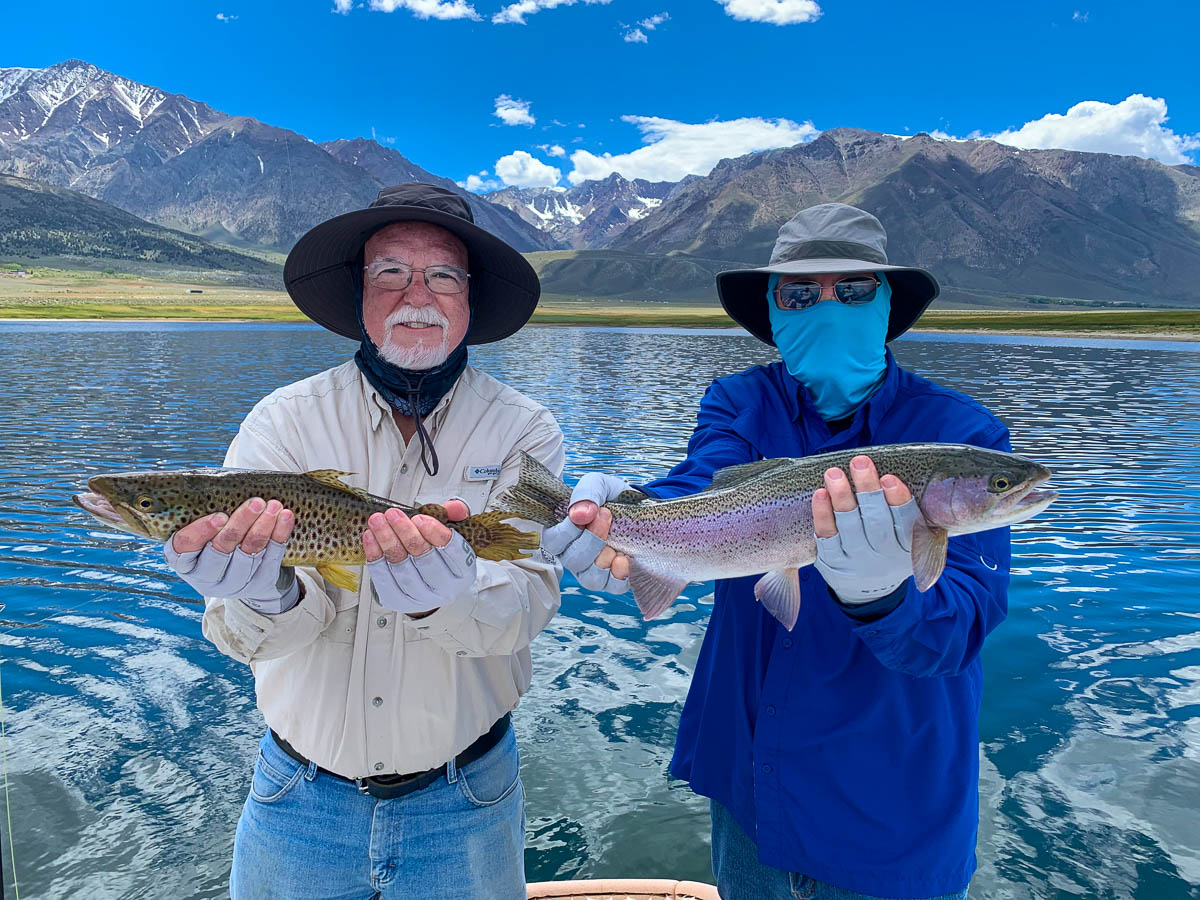 A pair of men wearing wide brim hats and holding two trout on a lake.