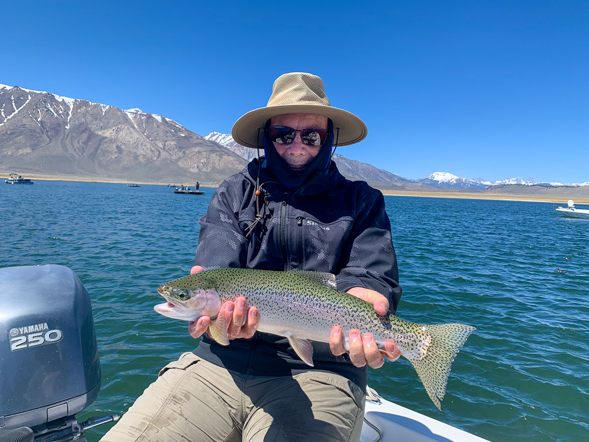 A fly fisherman with a wide-brim hat holding a massive cutthroat trout on a lake with snowy mountain tops behind him.