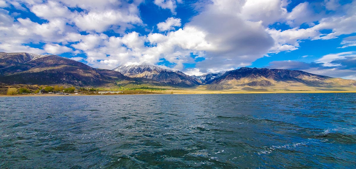 A mountain lake sitting against large mountains with snow on the tops and clouds above them.