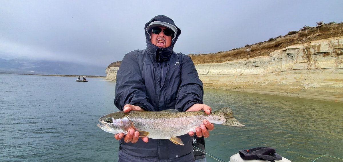 A hooded angler holding a rainbow trout on a mountain lake.