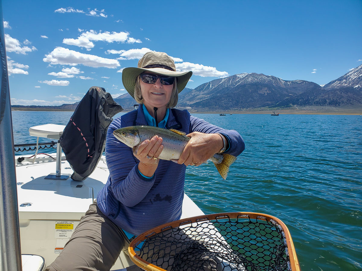 A female fly fisherman with a wide-brim hat holding a cutthroat trout on a lake.