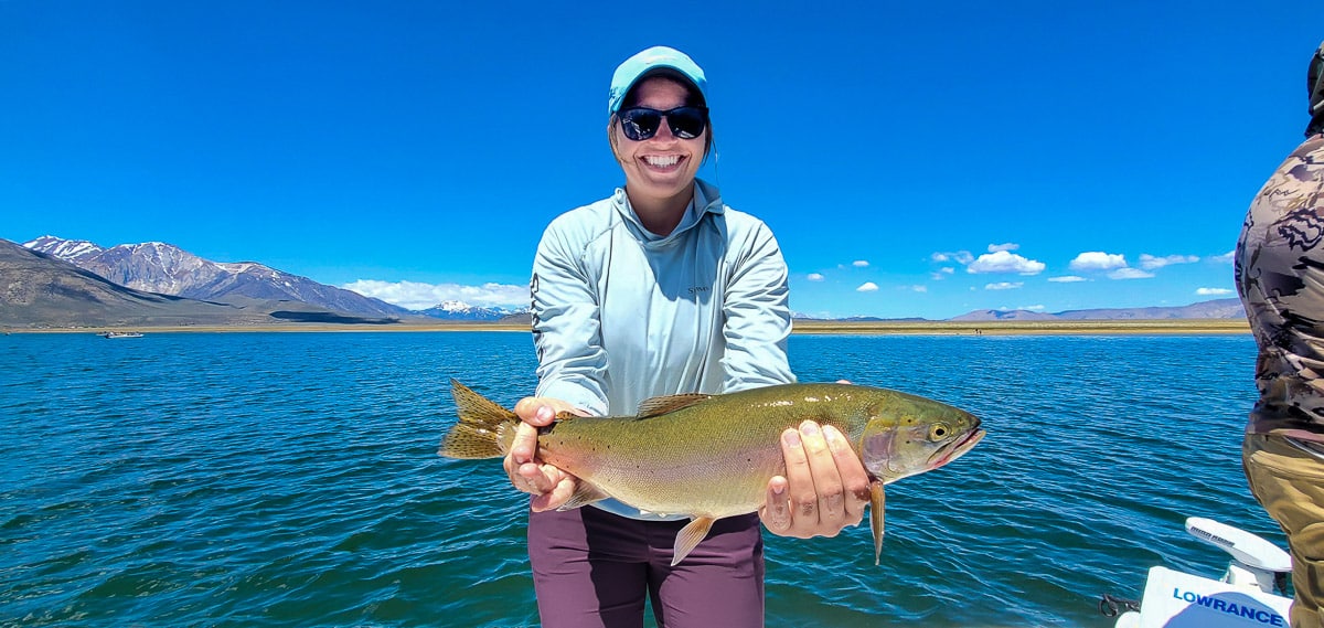 A female angler with a blue hat holding a large cutthroat trout on a lake.