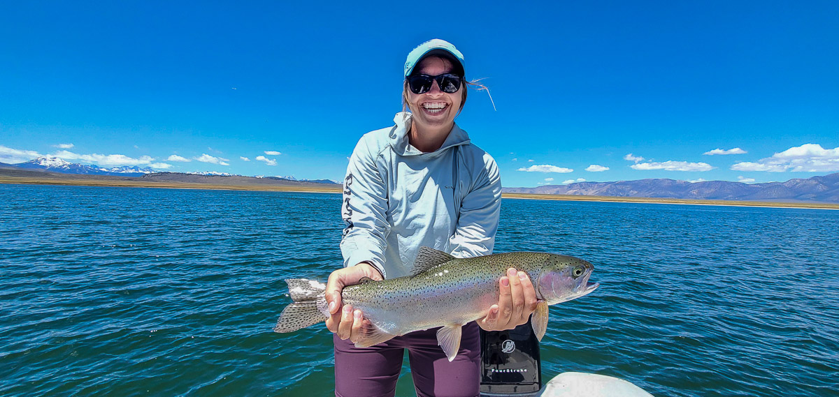 A female angler with a blue hat holding a large rainbow trout on a lake.