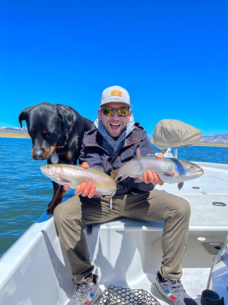 A young fly fisherman and a rottweiler dog inspecting a couple of rainbow and cutthroat trout while on a boat on a lake.