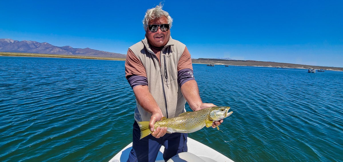 A fly fisherman holding his larger brown trout catch in a boat on a lake.