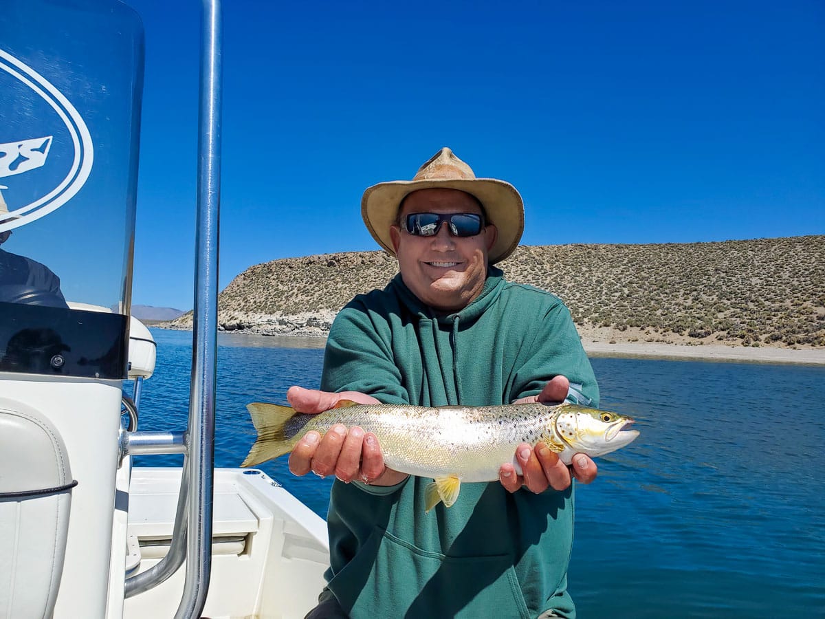 A fly fisherman with a wide-brim hat holding a cutthroat trout on a lake. 