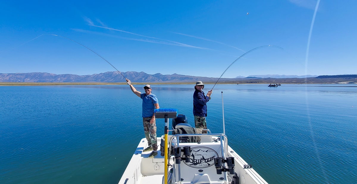 A pair of fly fishermen hooked up to trout and the same time in a boat on a mountain lake.