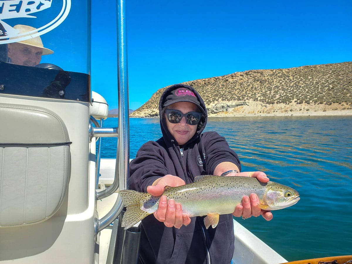 A female angler with a dark hoodie holding a large rainbow trout on a lake.