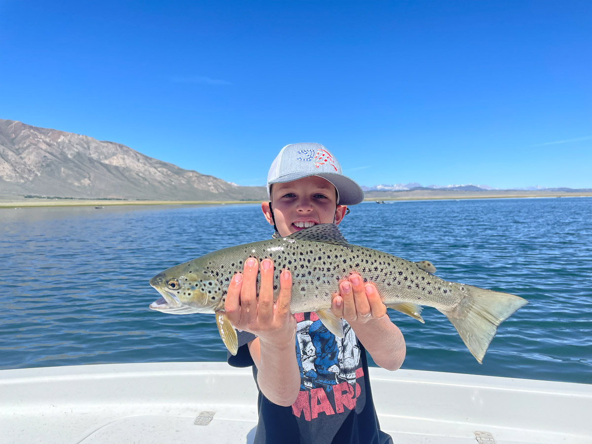 A young girl with a grey hat holding a large cutthroat trout in a boat on a lake.
