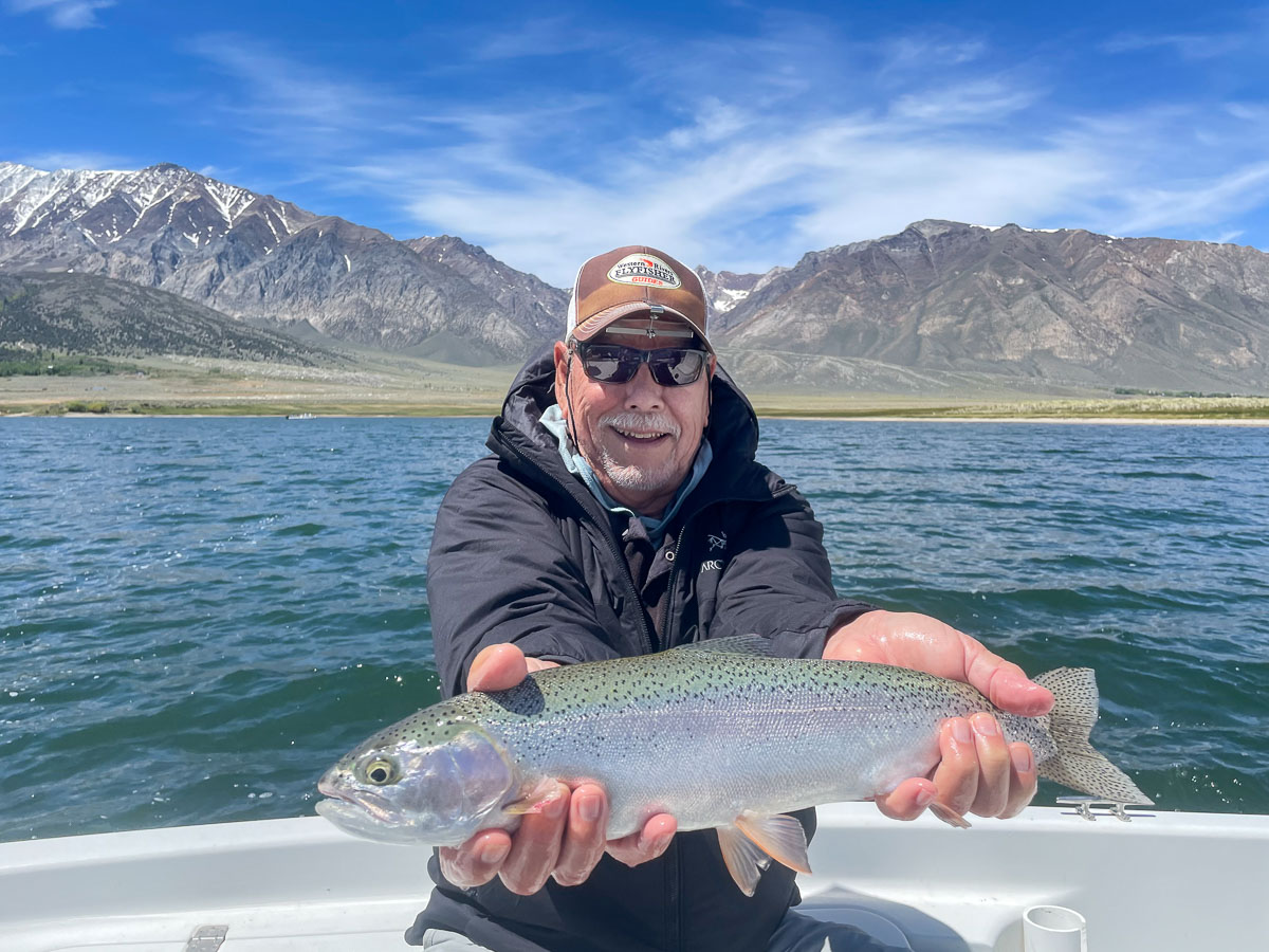 A smiling fly fisherman holding a rainbow trout on a lake in a boat.