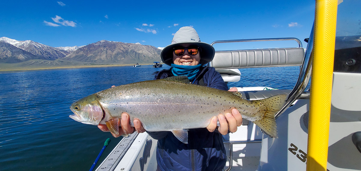A lady angler with a grey hat holding a giant cutthroat trout in a boat on a lake.