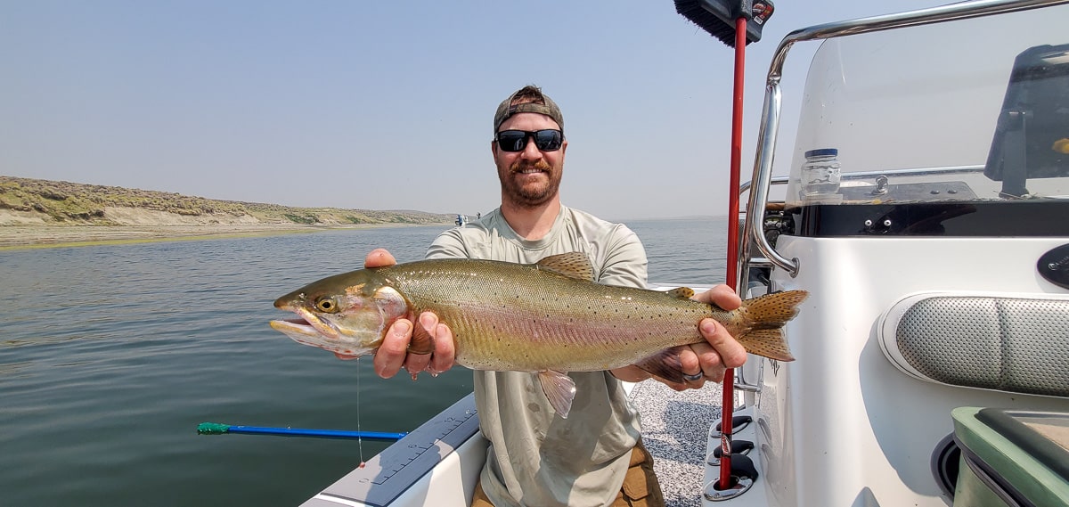 A smiling fly fisherman holding a cutthroat trout on a lake in a boat.