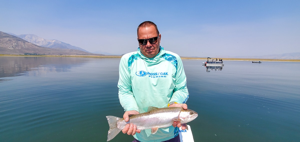 A smiling fly fisherman holding a rainbow trout on a lake in a boat.