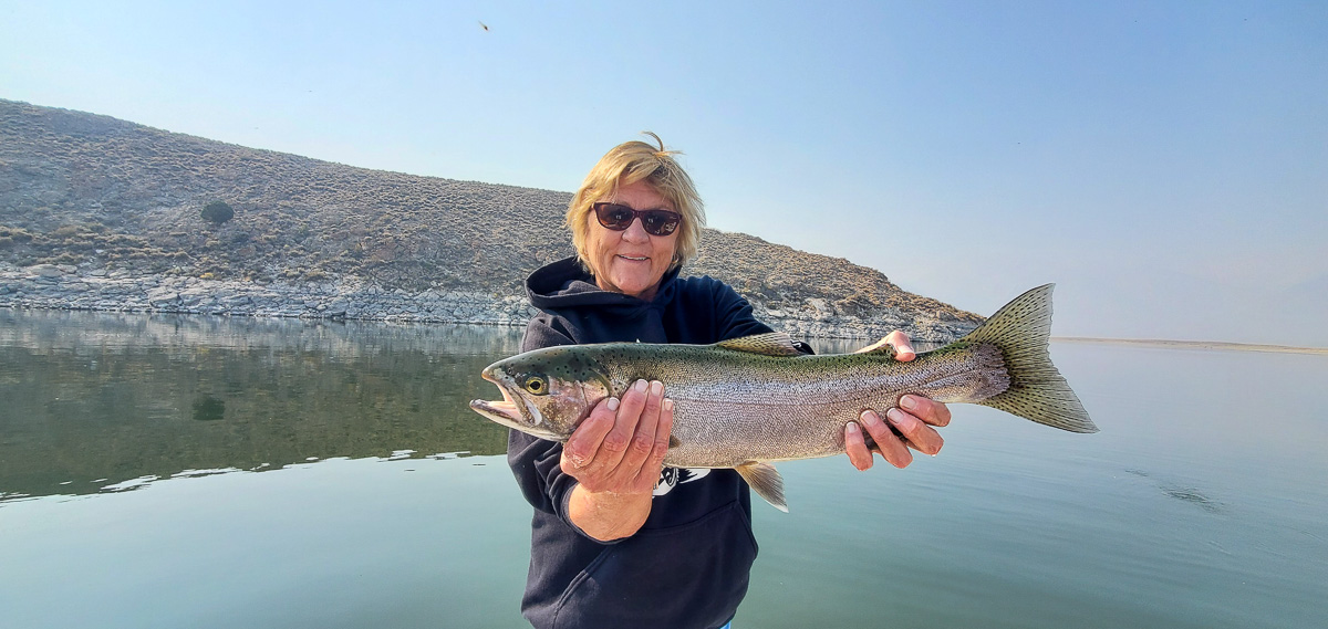 A smiling female fly fisherman holding a rainbow trout on a lake in a boat.