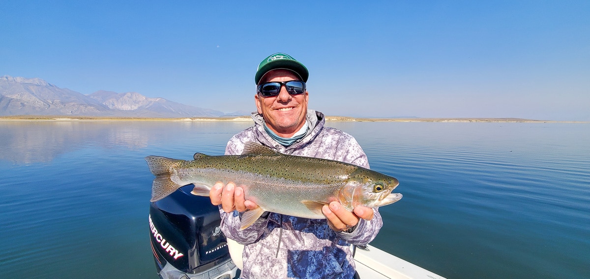 A smiling fly fisherman holding a rainbow trout on a lake in a boat.