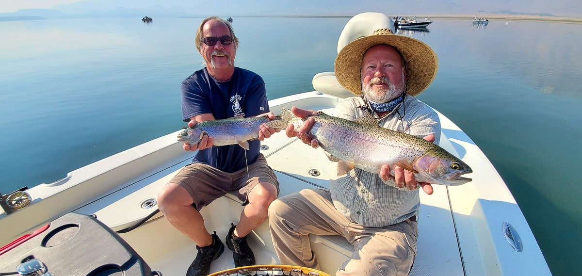 A pair of smiling fly fisherman holding 2 rainbow trout on a lake in a boat.