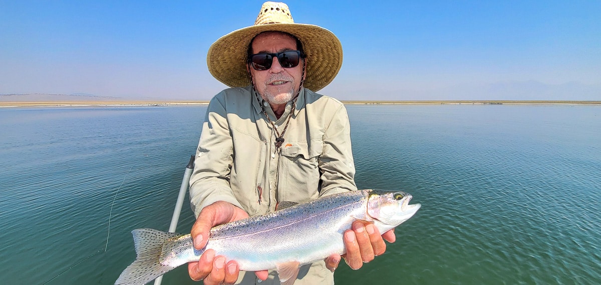 A smiling fly fisherman holding a rainbow trout on a lake in a boat.