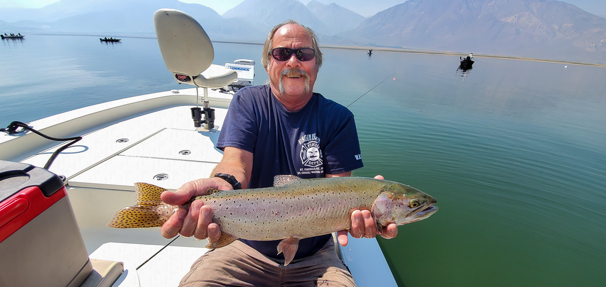 A smiling fly fisherman holding a cutthroat trout on a lake in a boat.