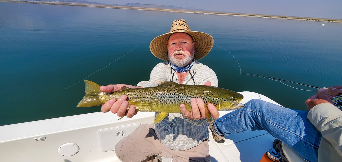 A smiling fly fisherman holding a brown trout on a lake in a boat.