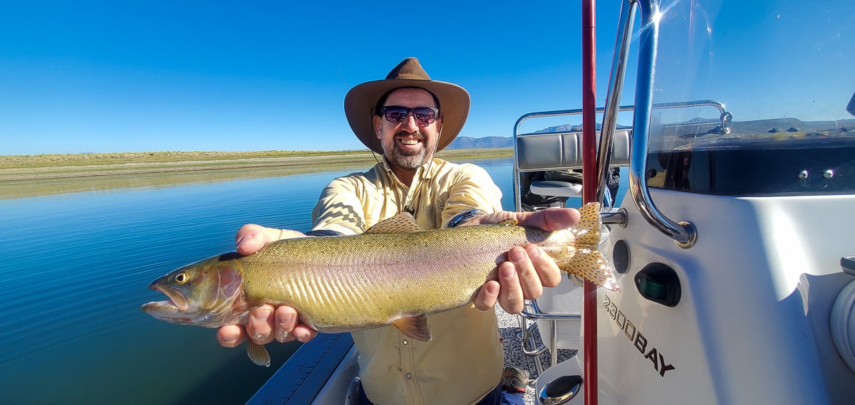 A smiling fly fisherman holding a cutthroat trout on a lake in a boat.