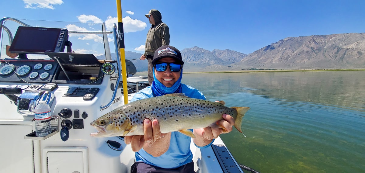 A smiling fly fisherman holding a rainbow trout on a lake in a boat.