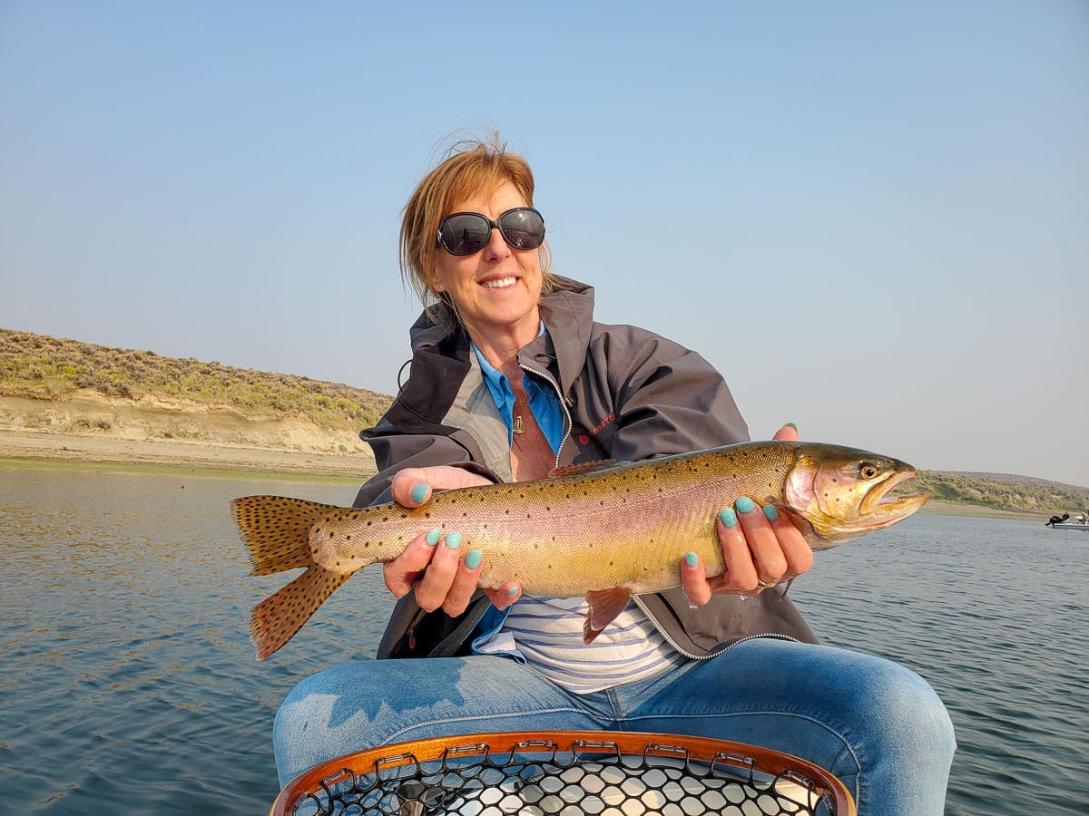 A female angler holding a large cutthroat trout on a lake.